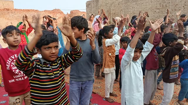 Children stand on a rug at a brick kiln with hands raised during a song