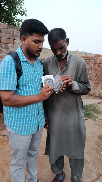 A young man shows an older man how to use a small audio player beside a brick wall