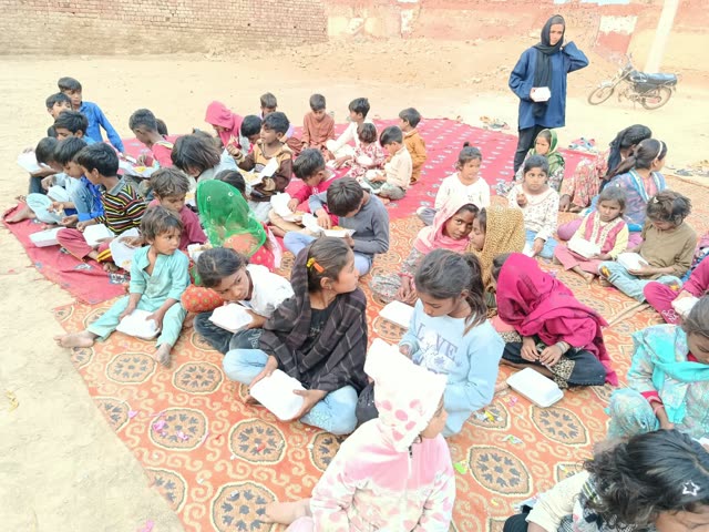 About forty children sit on rugs with boxed meals while a woman stands nearby holding a box