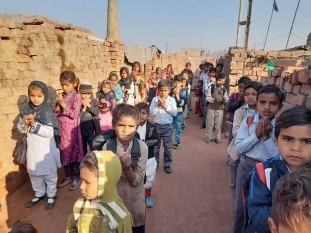 Children in school uniforms stand in rows with folded hands for prayer between brick walls