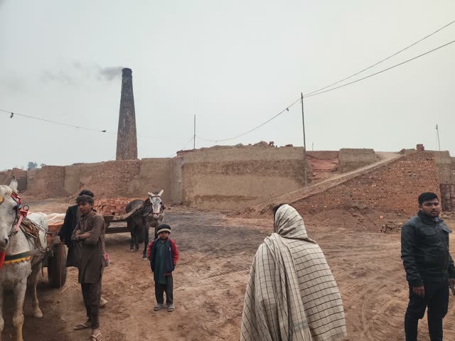 A brick kiln chimney rises behind donkey carts and people on bare ground