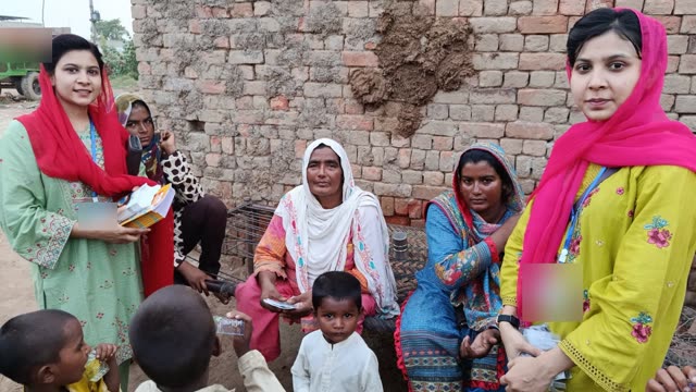 Two women share booklets and small devices with an older woman and children beside a brick wall