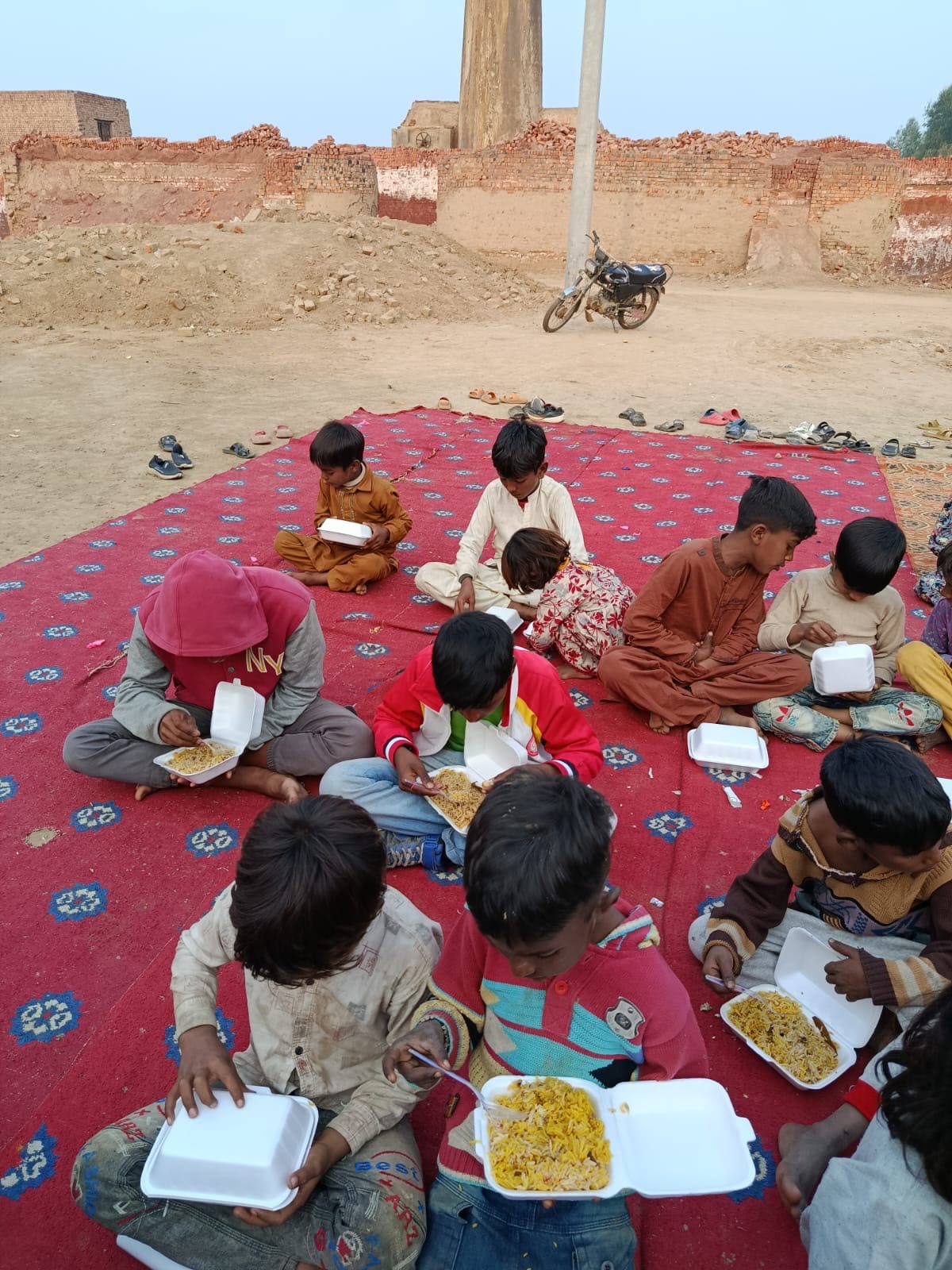 Children sitting on a red rug eating meals near brick walls and a tall chimney