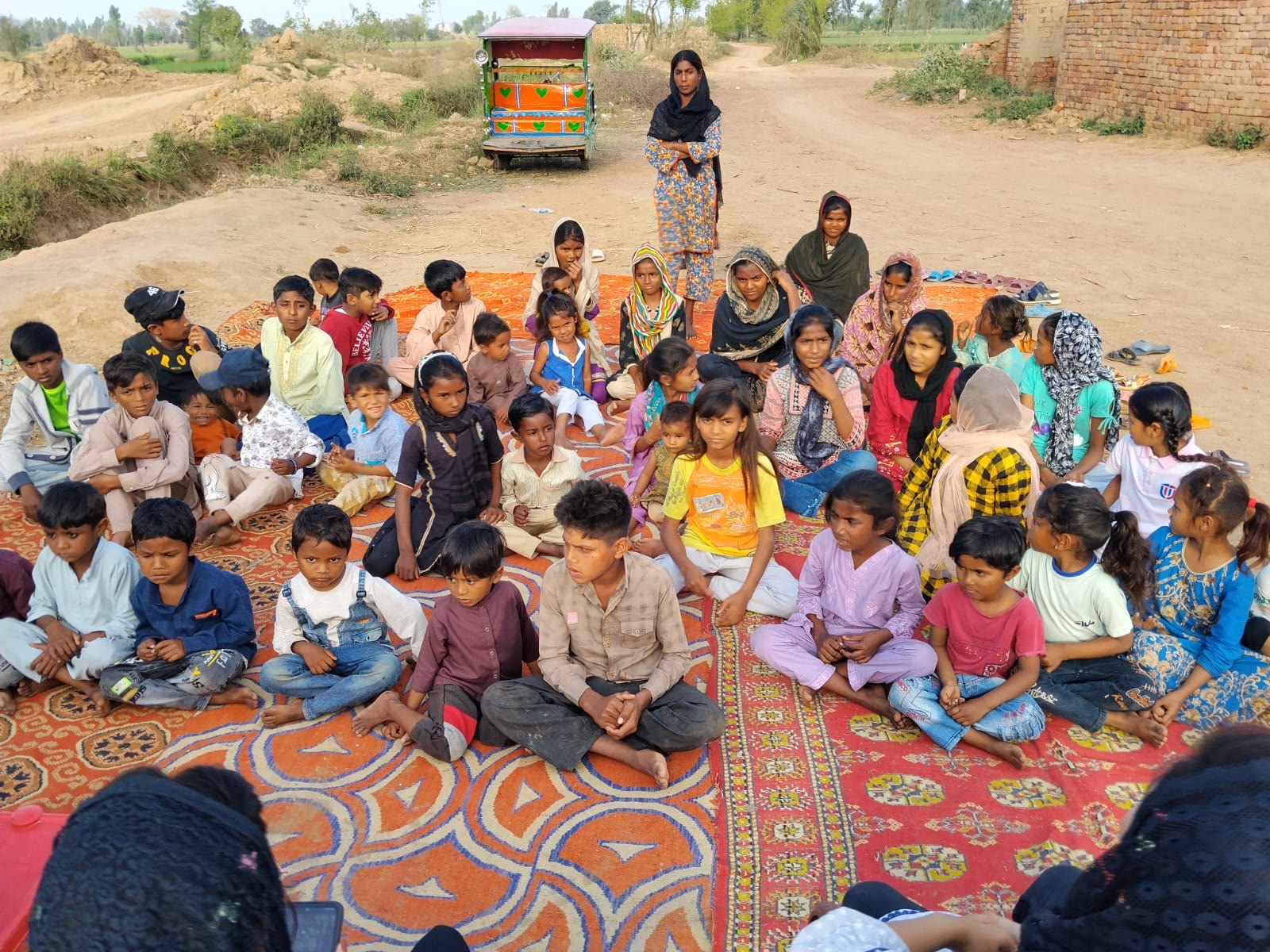 Large group of children seated on patterned rugs beside a rural road